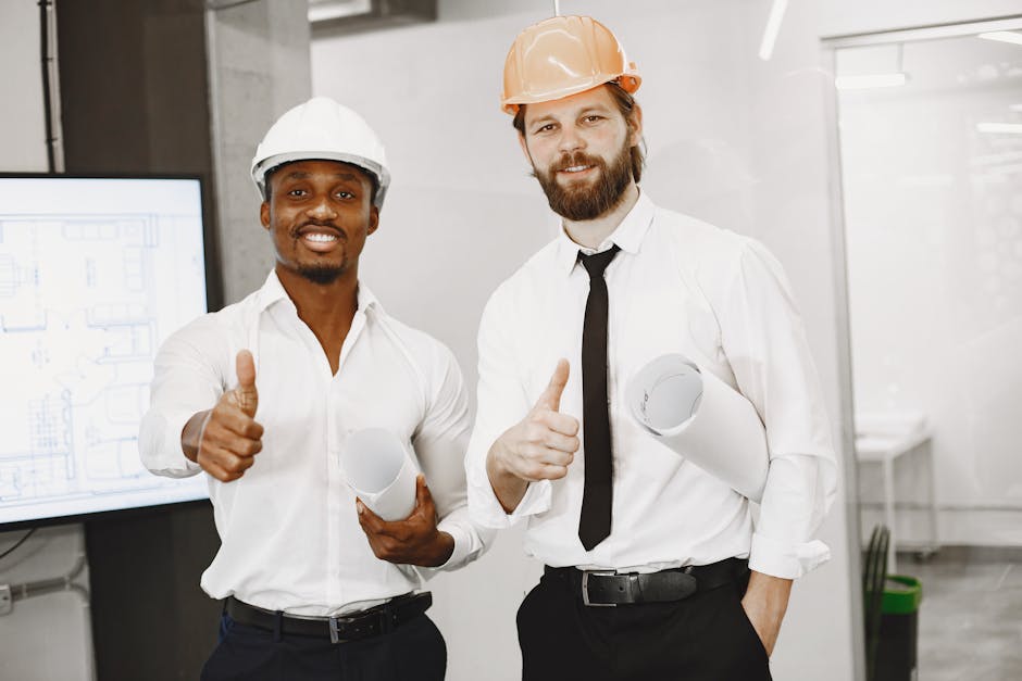 Two architects in hard hats give thumbs up in an office setting, holding blueprints and smiling confidently.